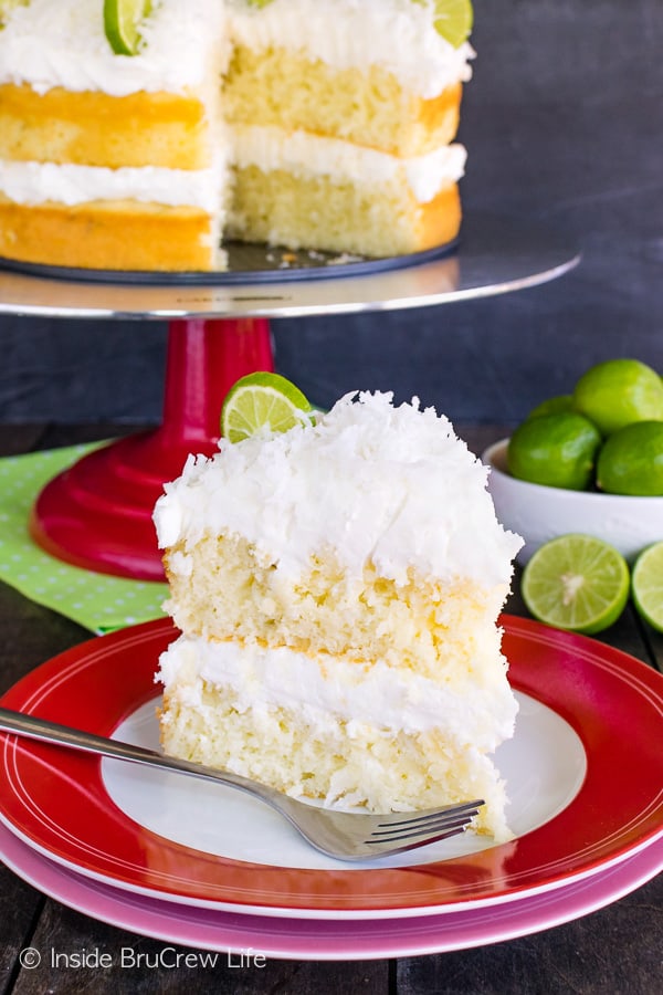 A slice of key lime cake topped with coconut frosting on a red and white plate with a cake plate behind it