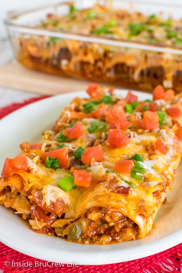 Two Monterey Chicken Enchiladas topped with green onions and tomatoes on a white plate with a pan of enchiladas behind it