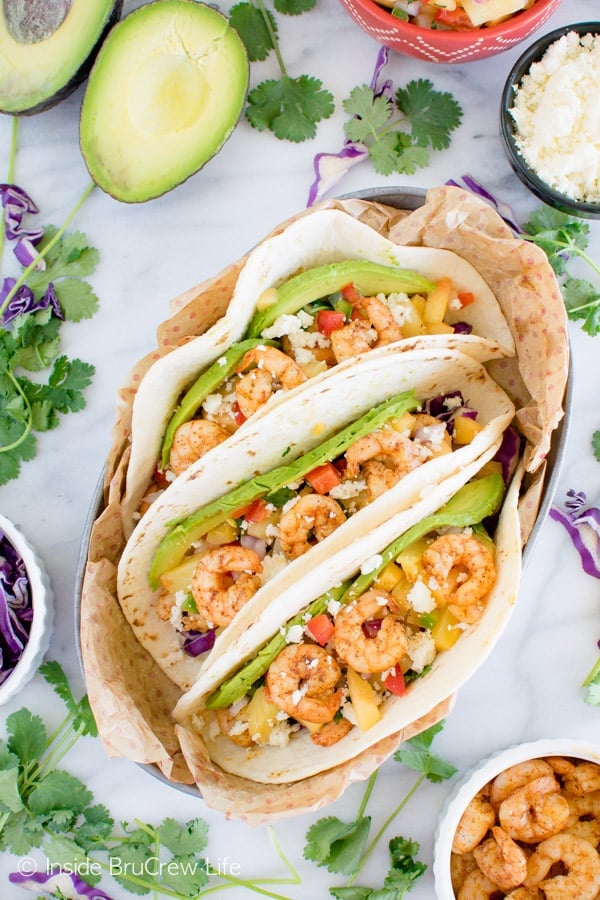Overhead picture of three shrimp tacos side by side in a dish with extra toppings all around it