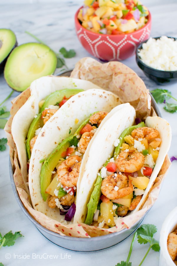 Close up of three tortilla shells in a dish loaded with shrimp, avocado slices, fruit salsa, and cabbage