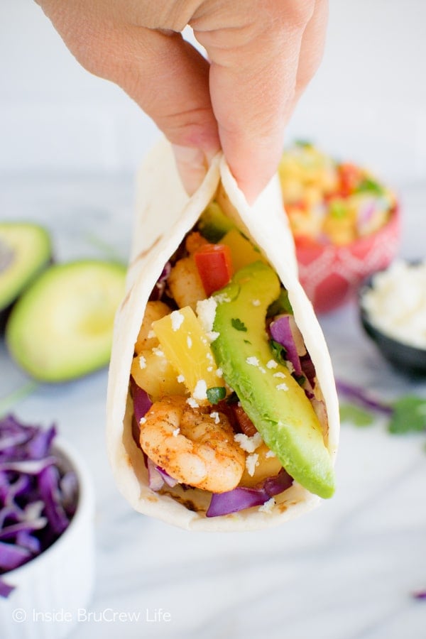 A close up picture of a hand holding a tortilla filled with shrimp, fruit salsa, and avocado slices up in the air