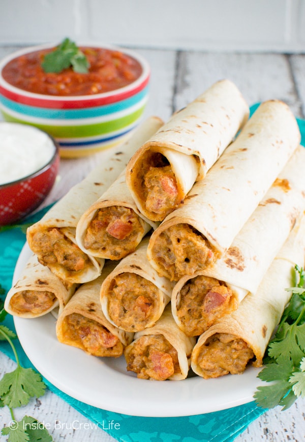 Taquitos stacked together on a white plate with salsa and sour cream in the background.