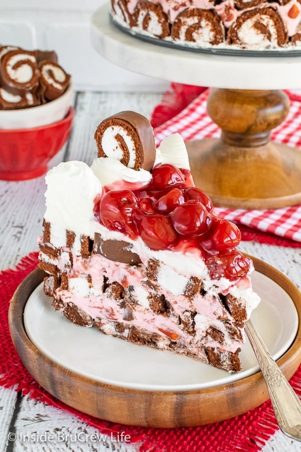 A slice of cherry swiss rolls cake on a white plate with the rest of the cake behind it