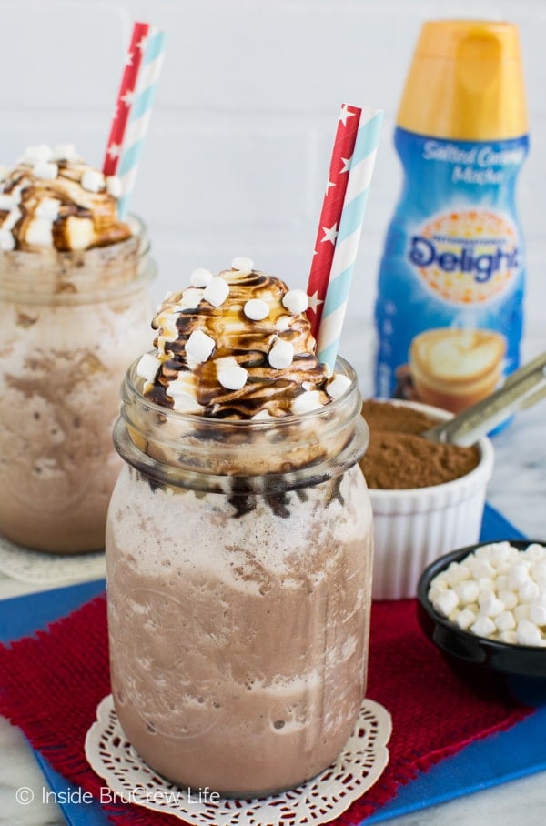 Two glasses filled with Frozen Hot Chocolate with bowls of hot chocolate mix and marshmallows behind them.