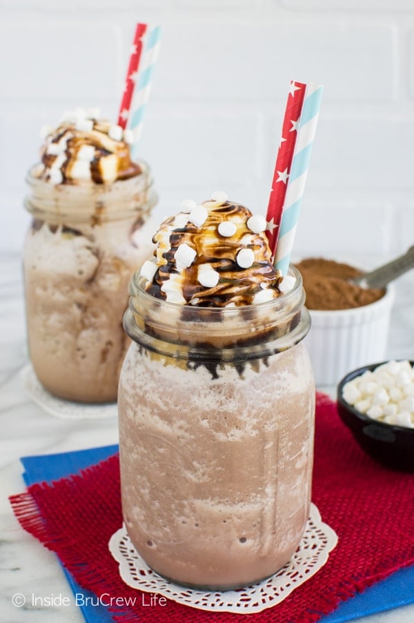 Two glasses filled with Frozen Hot Chocolate drizzled with caramel and chocolate with two straws in each.
