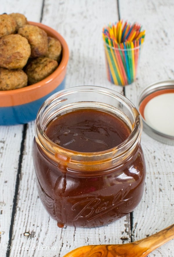 A jar of honey sriracha barbecue sauce with a bowl of baked meatballs behind it