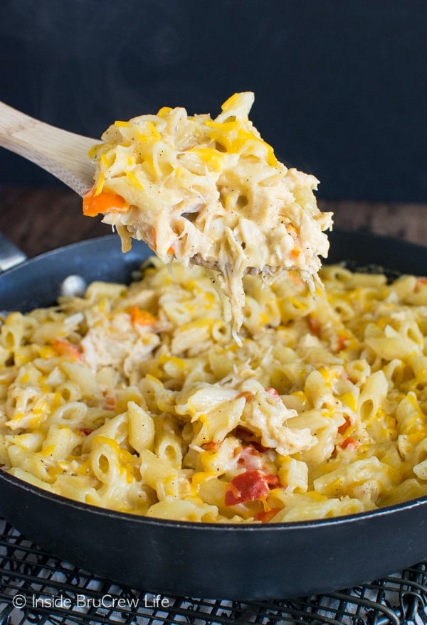 A skillet filled with cheesy chicken pasta with a wooden spoon lifting a big scoop out.