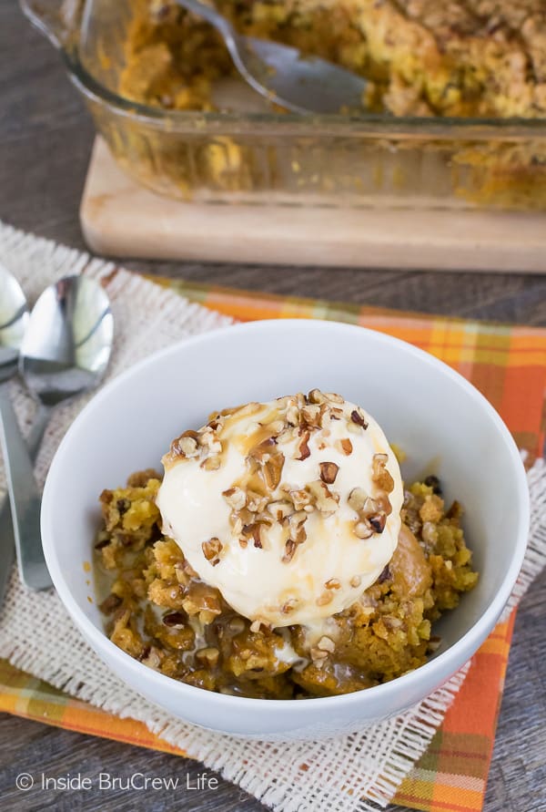 A white bowl filled with warm pumpkin cobbler topped with vanilla ice cream and pecans.