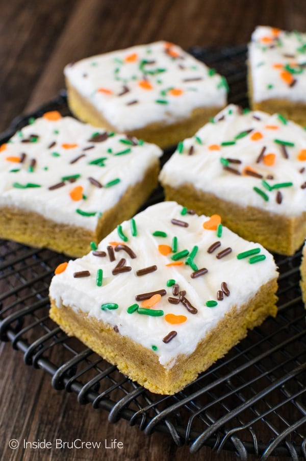A black wire rack with squares of frosted pumpkin sugar cookie bars with sprinkles on it