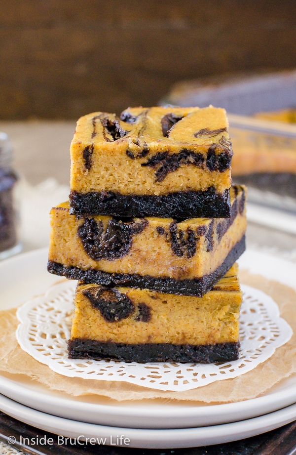 Three pumpkin cheesecake brownies stacked on top of each other on a white plate.
