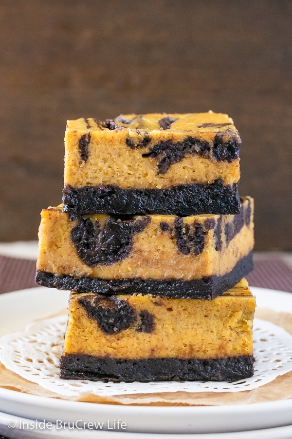 Three squares of pumpkin cheesecake brownies stacked on top of each other on a white plate.