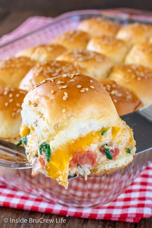 A pan with Cheesy Chicken Sliders in it with a metal spatula lifting one out showing the gooey filling on the side