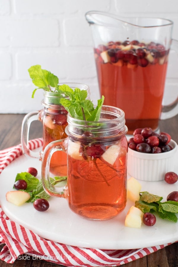 A white tray with two clear glasses filled with fruit punch, mint leaves, and fresh fruit with a pitcher of punch behind them