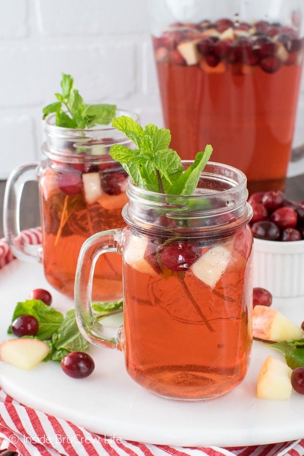 Two glasses filled with cranberry punch, mint leaves, and floating fruit on a white tray with a pitcher of punch behind it