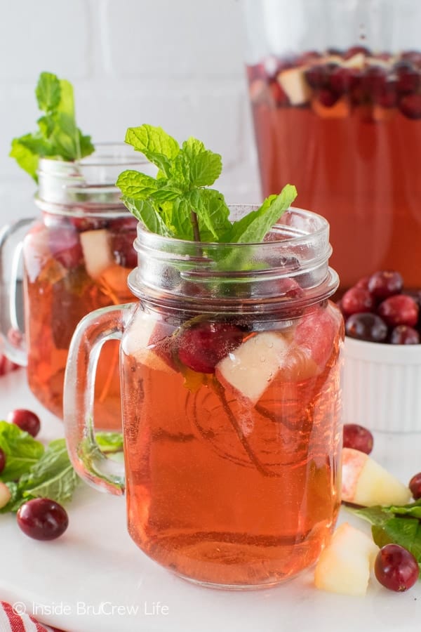 A clear glass filled with cranberry apple punch, mint leaves, and fresh fruit on a white tray with a pitcher of punch behind it