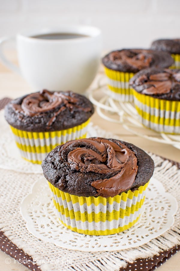 Two chocolate nutella banana muffins on white doilies with a tray of muffins behind them