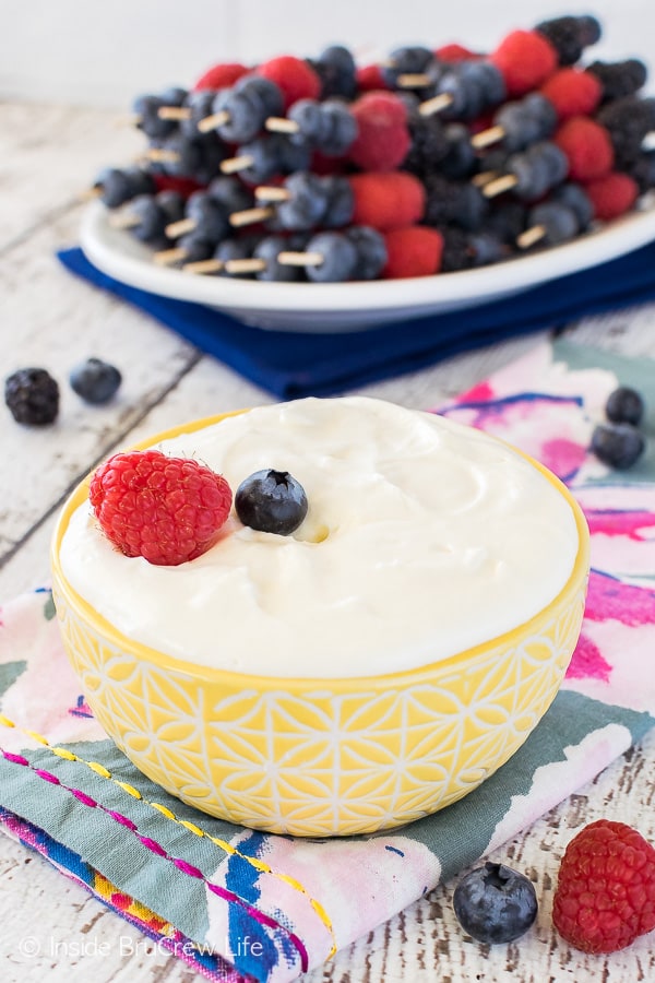 A yellow bowl filled with Lemon Cream Fruit Dip with a plate of fruit skewers behind it
