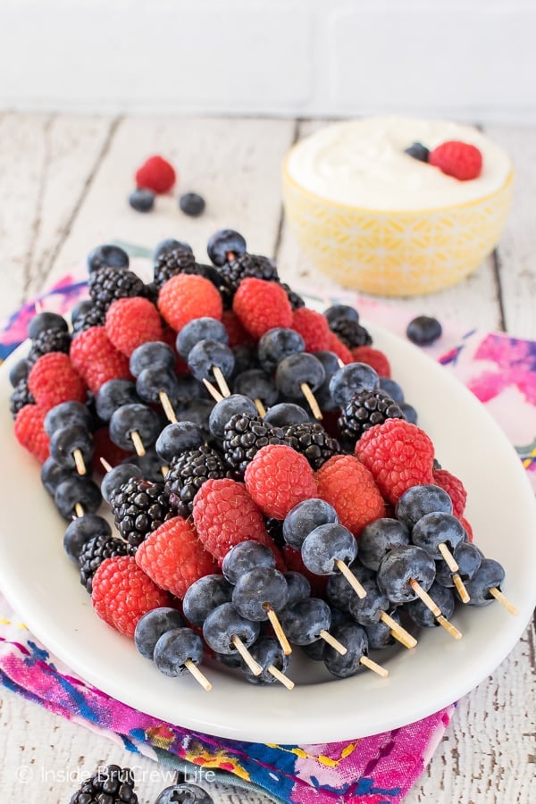 A white plate filled with fruit skewers and a bowl of lemon cream fruit dip behind it