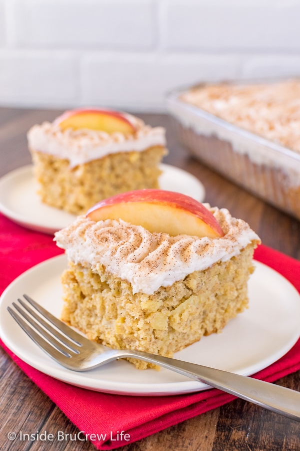 Two white plates on a red towel with squares of snickerdoodle apple cake with vanilla frosting and apple slices.