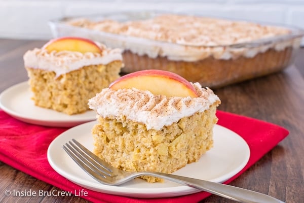 Two white plates on a red towel with squares of snickerdoodle apple cake with cinnamon frosting and apple slices.