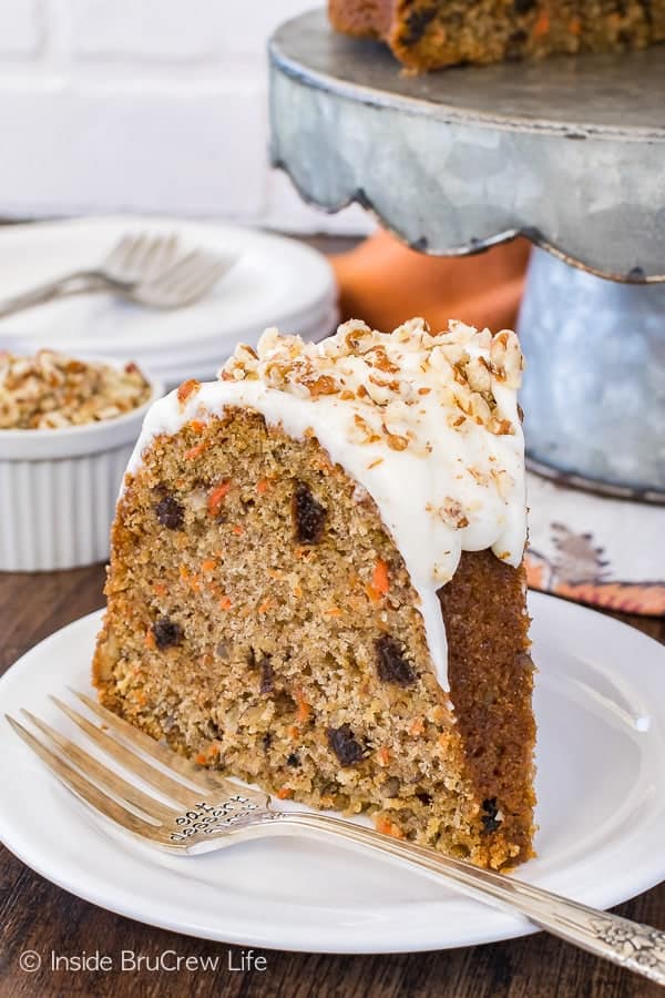 A slice of carrot bundt cake filled with raisins, spices, and carrots topped with a white frosting and pecans on a white plate.