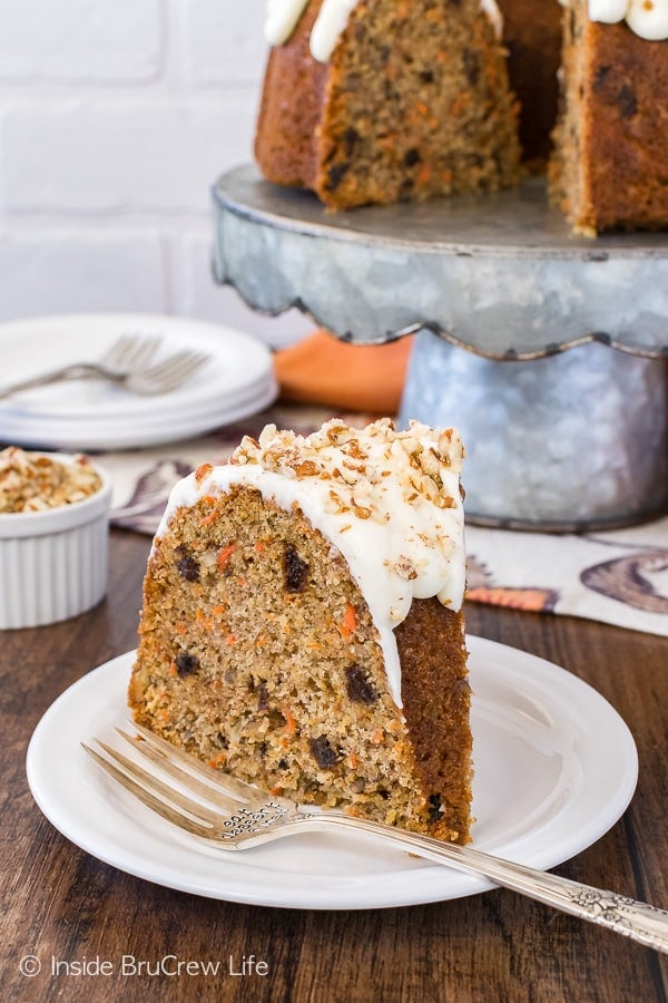 A slice of carrot bundt cake filled with raisins, spices, and carrots topped with a white frosting and pecans on a white plate.