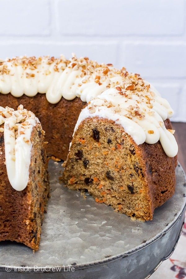 A carrot bundt cake filled with raisins, spices, and carrots topped with a white frosting and pecans on a metal cake plate.