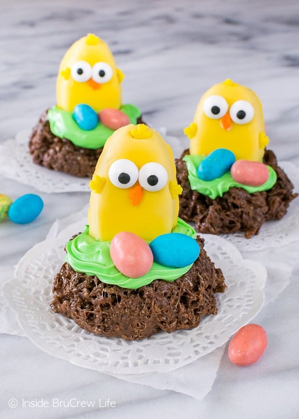 A peanut butter cup decorated as a small chicken on a chocolate nest with 2 colorful eggs by it.