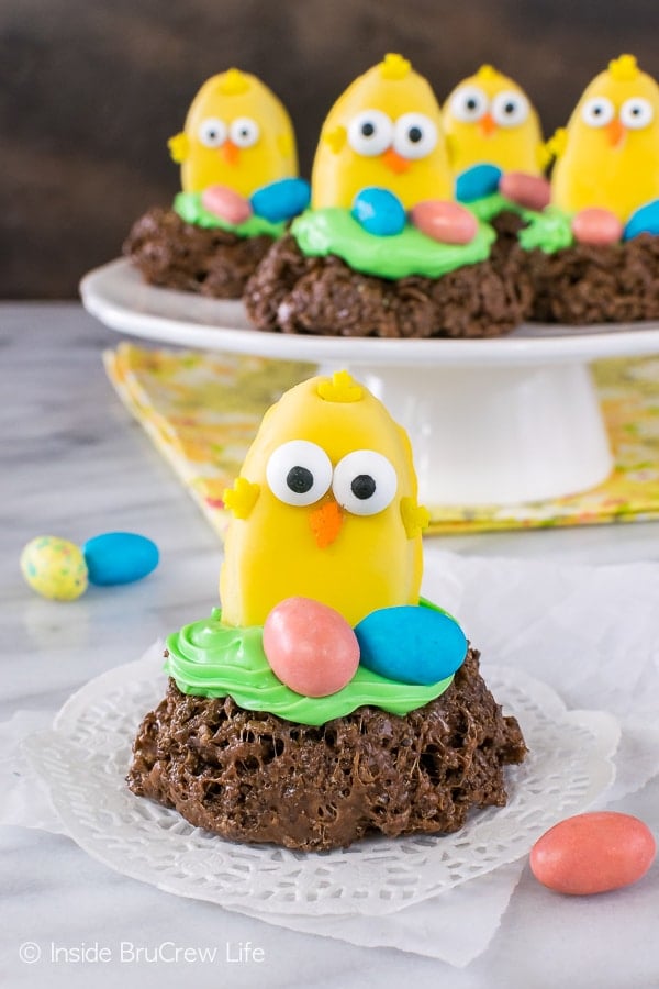 A peanut butter cup decorated as a small chicken on a chocolate nest with 2 colorful eggs by it.