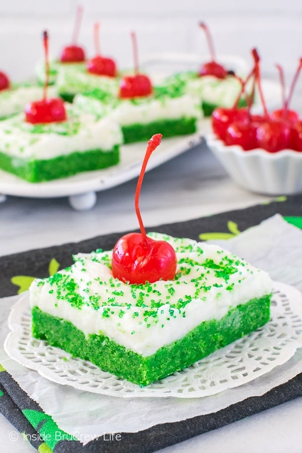 A square of Shamrock Shake Sugar Cookie Bars with white frosting, green sugar, and a maraschino cherry on a white doily with more bars behind it