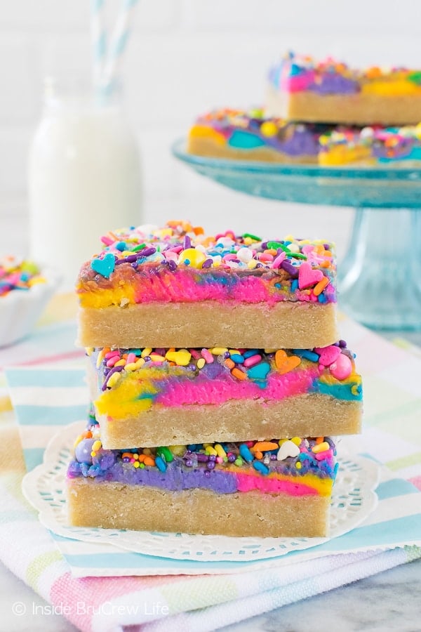 Three frosted sugar cookie bars with unicorn sprinkles stacked on top of each other with more bars behind it