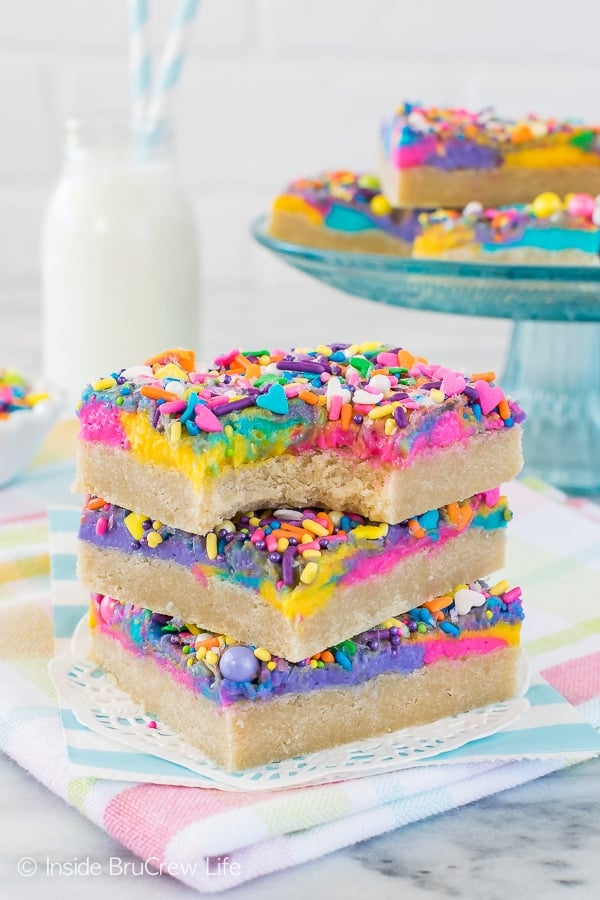 Three sugar cookie bars frosted with colorful vanilla frosting and sprinkles stacked on top of each other with a bite out of the top one