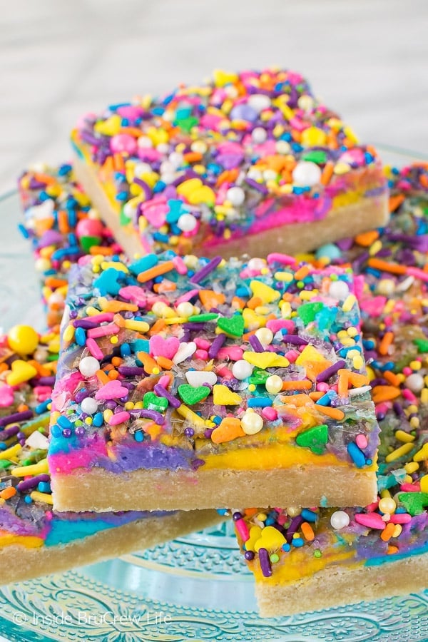 Sugar cookie bars frosted with colorful vanilla frosting and unicorn sprinkles stacked on a blue plate