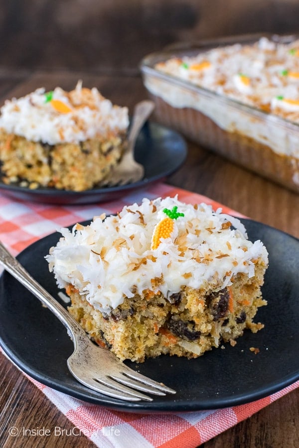 A piece of carrot cake with white frosting topped with toasted coconut on a black plate.