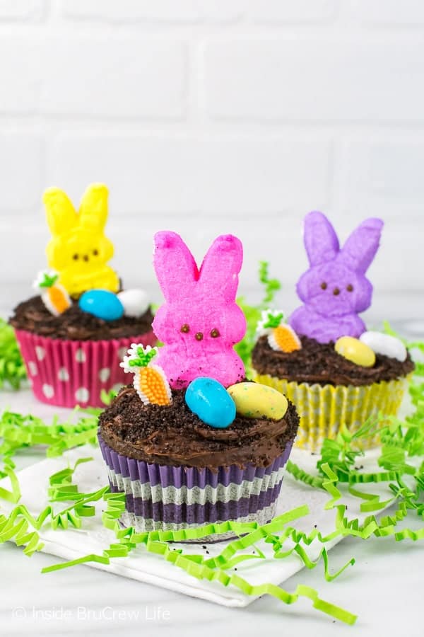 3 chocolate cupcakes topped with malt eggs, a candy carrot, and marshmallow bunnies.