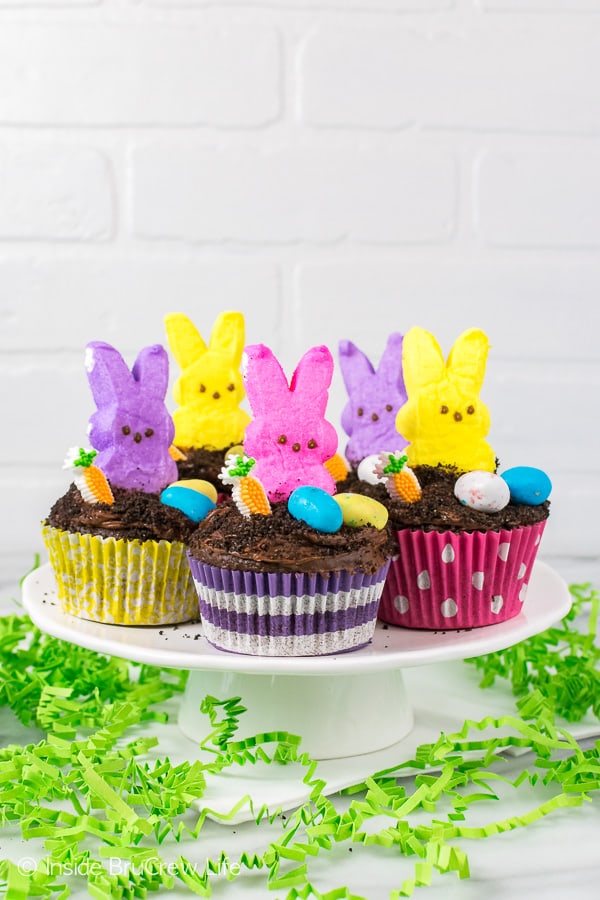 A white cake plate full of chocolate cupcakes topped with malt eggs, a candy carrot, and marshmallow bunnies.