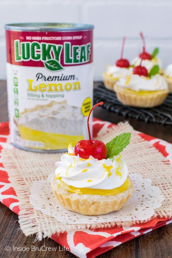 Mini lemon cheesecake pies topped with cherries on a white doily with a can of pie filling in the background.