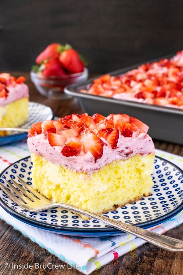 A slice of Lemon Strawberry Poke Cake on a blue and white plate with the cake behind it.