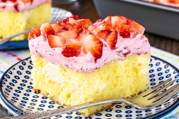 A piece of lemon poke cake topped with strawberry whipped cream and fresh strawberries on a white and blue plate.