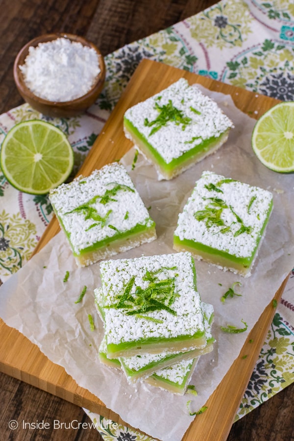 An overhead picture of a wooden cutting board with key lime bars with powdered sugar on them scattered on it.