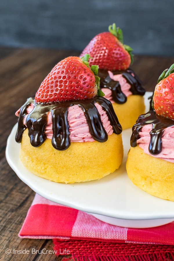 Three strawberry mousse shortcakes with chocolate drizzles and strawberries on a white cake plate.