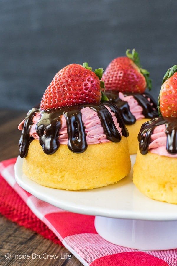 A white cake plate with three strawberry mousse cakes topped with chocolate drizzles and fresh strawberries. 