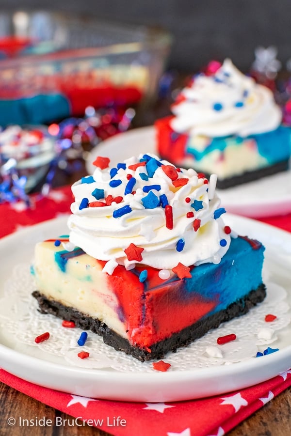 A white plate with a square of red white and blue cheesecake bar topped with whipped cream and sprinkles