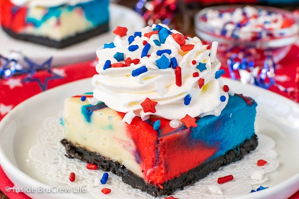A white plate on a red towel with a square of red white and blue cheesecake bars topped with Cool Whip and patriotic sprinkles