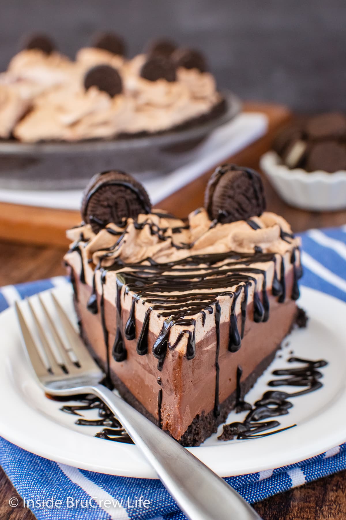 A white plate with a slice of chocolate pie topped with hot fudge and Oreos.