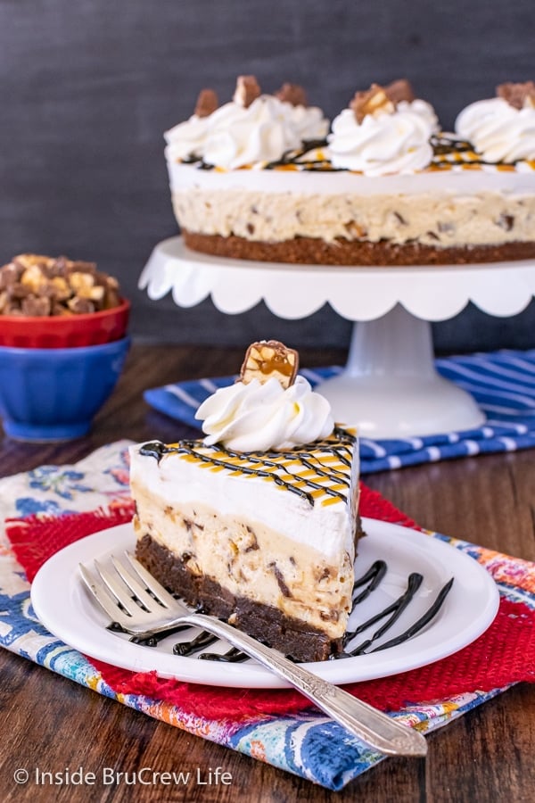 A white plate on a red towel with a slice of no bake snickers cheesecake with a brownie crust on it