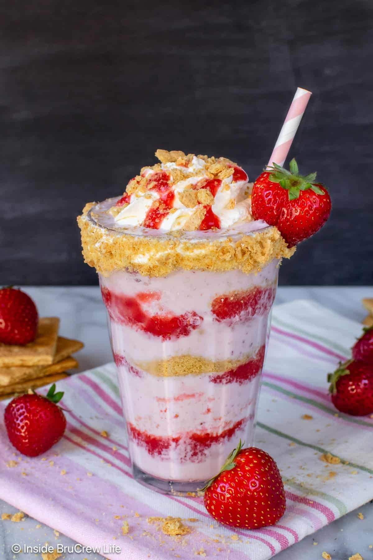 A clear glass filled with strawberry cheesecake milkshake with layers of puree and graham cracker crumbs.