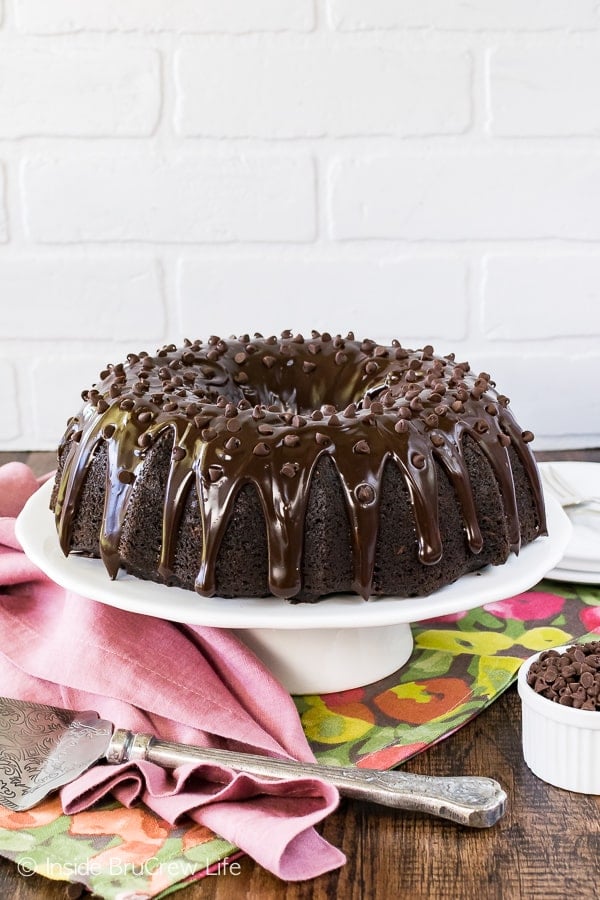 A white cake plate with a full bundt cake with chocolate glaze on it.