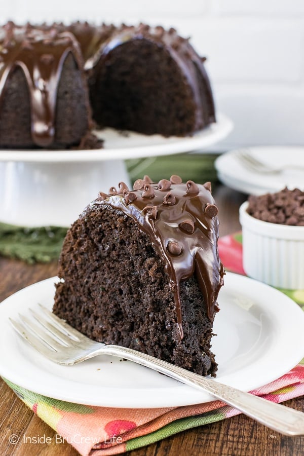 A white plate with a fork and a slice of chocolate zucchini cake on it and more cake behind it.