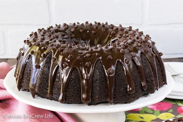 A white cake plate with a full bundt cake with chocolate glaze.